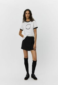 Vitt t-shirt med svart ringerkrage och tryck av "HOTEL IMAGINE", kombinerat med en svart mini wrap kjol och knähöga svarta strumpor.
