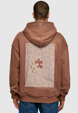 Brun oversized hettegenser med et stort puslespill-design i beige og lysebrune nyanser på baksiden, laget av et teksturert og behagelig stoff.
