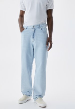 Mand iført lyseblå løse jeans, hvid t-shirt med korte ærmer og hvide slip-on sko, stående med den ene hånd i lommen.