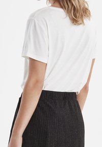Biała bawełniana koszulka z krótkim rękawem, widok z tyłu, w połączeniu z czarnymi spodniami plisowanymi z subtelnymi złotymi paskami.