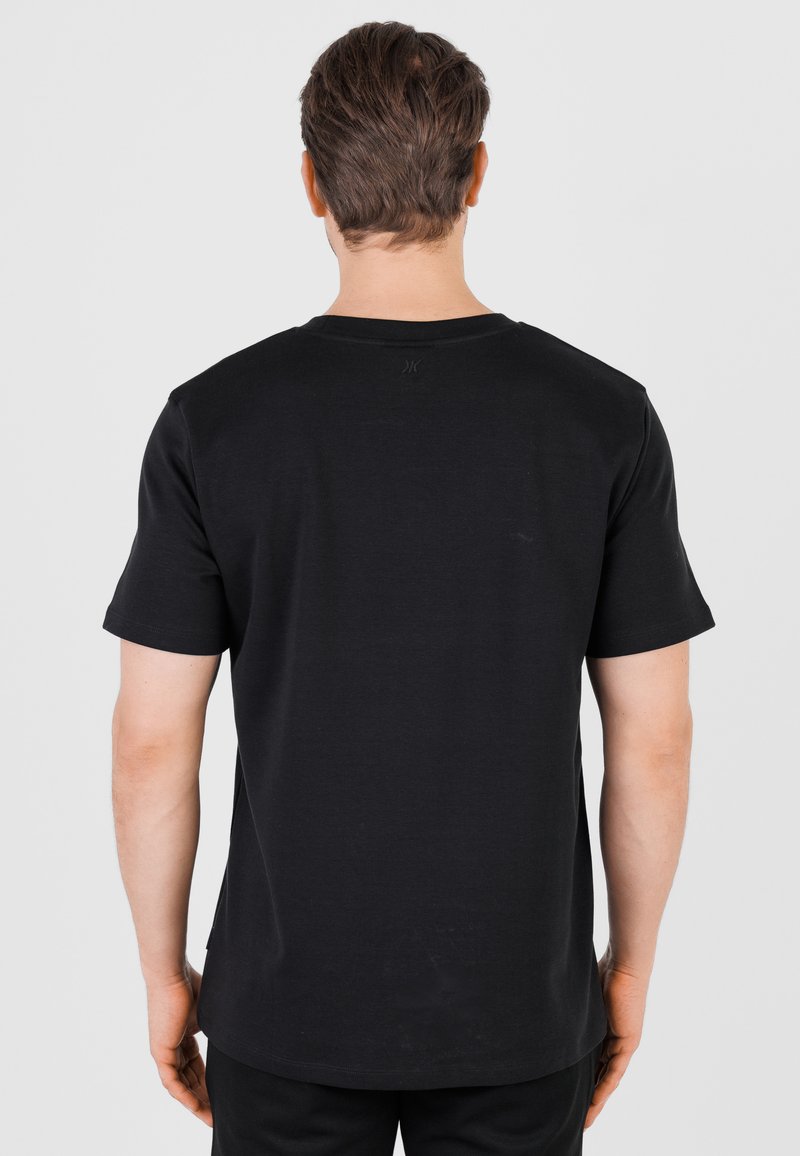 Homme aux cheveux bruns courts portant un t-shirt noir uni à manches courtes, vu de dos sur un fond gris clair uni.