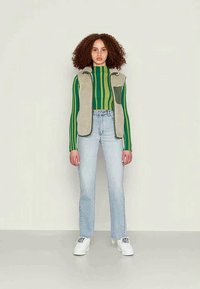 Mulher com cabelo encaracolado, vestindo uma blusa de mangas longas às riscas verde e amarela, um colete de fleece bege, jeans claros e ténis brancos, de pé contra uma parede simples.