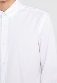 Valkoinen napitettu paita, jossa on kaulus ja pitkät hihat, henkilön päällä, kuvattu kaulasta vyötärölle.