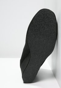 Chaussure noire avec une semelle en caoutchouc texturé, présentant une forme arrondie et une surface supérieure lisse. Aucun matériel ou accent visible.