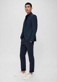 Dunkelblazer über einem schwarzen Hemd, mit marineblauen Hosen und weißen Turnschuhen. Maßgeschneiderte Passform, glatte Textur und dezente Muster.