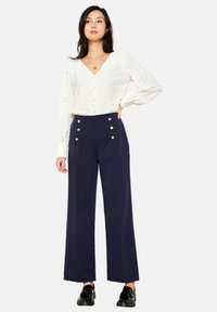 Blusa bianca in pizzo con bottoni e maniche lunghe, abbinata a pantaloni navy a vita alta con bottoni dorati. Scarpe nere completano il look.