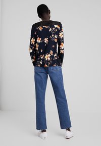 Blusa floreale a maniche lunghe in navy, con fiori arancioni e bianchi; dettagli in pizzo sul retro. Abbinata a jeans a gamba larga blu e scarpe bianche.