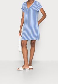 Blårandig kortärmad klänning med V-ringning, med vertikala vita ränder, avslappnad passform och fickor, parat med vita platta sandaler.