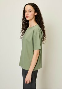 Olīvzaļš kokvilnas T-krekls ar noapaļotu kakla izgriezumu un īsām piedurknēm, piedāvā atbrīvotu piegriezumu un teksturētu audumu, apvienots ar tumšām biksēm.