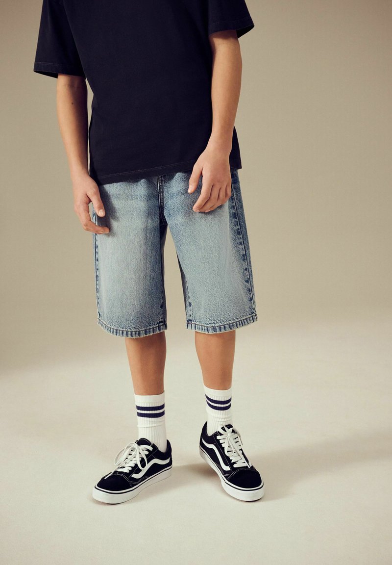 Personne portant un t-shirt noir, un short en jean bleu clair, des chaussettes blanches avec des rayures noires et des baskets noires avec des lacets blancs, debout sur un fond uni.