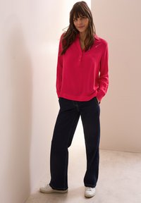 Fuchsia Langarmhemd mit Kragen und Knopfleiste, kombiniert mit weit geschnittenen dunkelblauen Jeans und weißen Sneakern vor einem hellen Hintergrund.