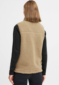 Gilet en polaire avec un corps texturé de couleur beige et des manches en tricot noires, avec un col montant et un ourlet droit, conçu pour être superposé.
