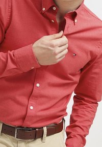 Rotes Hemd mit Knopfleiste und Kragen, in taillierter Passform, mit einem kleinen Logo auf der Brust. Der Stoff wirkt glatt mit subtiler Textur.