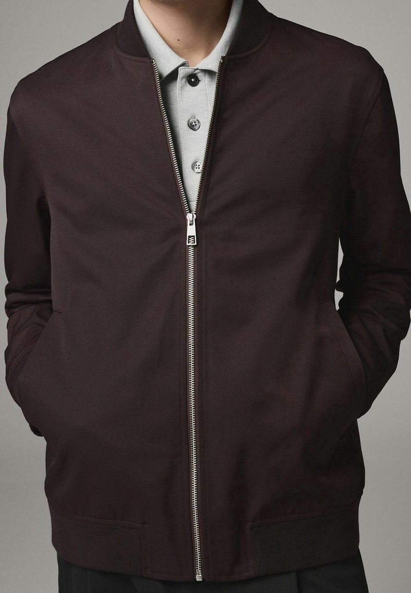 Homme portant une veste zippée marron foncé sur un polo gris clair, les mains dans les poches de la veste, debout devant un fond gris uni.