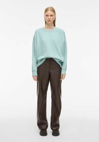 Donna con capelli biondi lisci indossa un felpa oversized di colore azzurro chiaro, pantaloni in pelle marrone scuro, scarpe marrone scuro e orecchini a cerchio in argento.