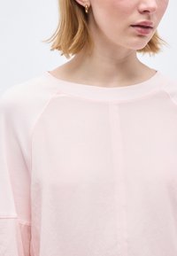 Νεαρή γυναίκα με κοντά ξανθά μαλλιά φοράει ροζ ανοιχτό τοπ και χρυσούς κρίκους σκουλαρίκια, κοιτάζοντας ελαφρώς πλάγια, μπροστά σε απλό φόντο.