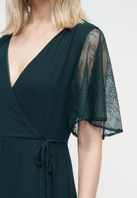 Γυναίκα που φοράει σκούρο πράσινο φόρεμα τύπου wrap με διάφανα μανίκια από δαντέλα και ζώνη στη μέση, εμφανιζόμενη από τον ώμο έως τη μέση.