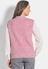 Frau mit langen grauen Haaren trägt eine ärmellose rosa Strickweste über einem weißen Langarmshirt und grauen Hosen, mit dem Rücken zur Kamera gewandt.