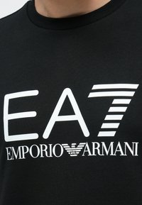 Közeli kép fekete pólóról, amelyen nagyméretű fehér "EA7" és kisebb "Emporio Armani" felirat, illetve logó látható a mellrészen.