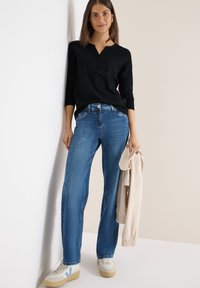 Musta pitkähihainen paita, jossa on kolme nappia, siniset farkut, joissa on leveät lahkeet, valkoiset sneakersit sinisillä yksityiskohdilla, pitäen vaaleanbeigeä takkia.