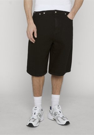 Mand iført sorte knælange shorts, hvide sokker og hvide New Balance-sneakers, stående med den ene hånd i lommen.