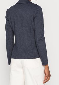 Námořnický blazer s texturovanou tkaninou, strukturovaným límcem, dlouhými rukávy a nenápadným švem, stylizovaný přes světle zbarvené kalhoty.