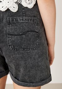 Detalles de un bolsillo trasero en unos pantalones cortos de mezclilla negros desgastados, combinados con una blusa blanca de ganchillo, mostrando parte del brazo de una persona.