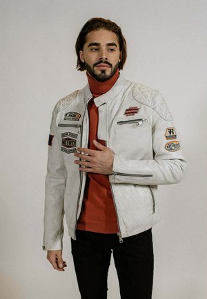 Homme barbu portant une veste de course blanche avec des écussons sur un col roulé rouille, debout devant un fond clair uni.