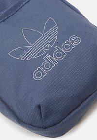 Blå texturerad påse med en upphöjd, vit Adidas-logotyp och tre horisontella ränder. Har en rundad form och en jämn yta.