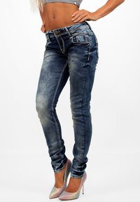 Jean slim-fit en un lavado azul desgastado con costuras en contraste, que presenta bolsillos y un cierre frontal con botón, acompañados de tacones altos brillantes.