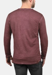 Mand i en langærmet maroon skjorte og mørkegrå jeans, vist fra ryggen mod en ensfarvet hvid baggrund.