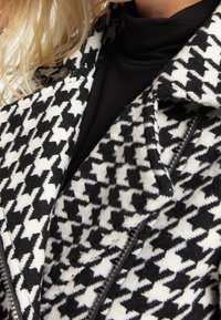 Abrigo de patrón de espiga en blanco y negro con tejido texturizado, cuello con muesca y detalle de cremallera en el costado.