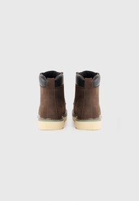 Une paire de bottines en cuir marron avec des cols rembourrés noirs et des semelles beiges, vues de l'arrière sur un fond blanc.