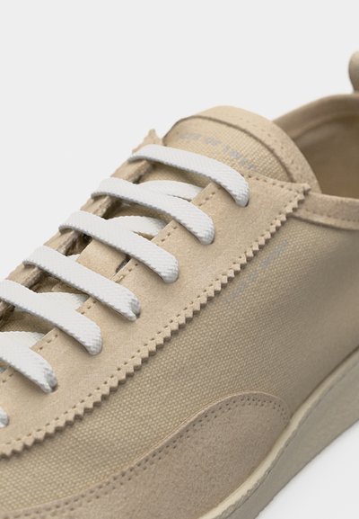 Smėlio spalvos sportbačiai su baltais raišteliais, drobinis ir zomšinis viršus, zigzago siuvimo detalė ir tekstūruota balta guminė padas.