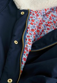 Jachetă bleumarin cu fermoar auriu; căptușită cu imprimeu floral în roșu, albastru și alb; guler moale din fleece crem; textură exterioară fină.