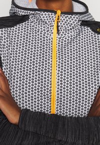 Persoon in een geblokte ritsjack met een gele rits en zwarte mouwen, met de armen over de borst gekruist.