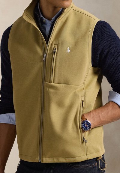 Khakinvärinen fleece-liivi vetoketjulla edessä, sivutaskuilla ja kirjailulla logolla. Rakenteelliset laivastonsiniset hihat ja alla kerroksittain sininen paita.