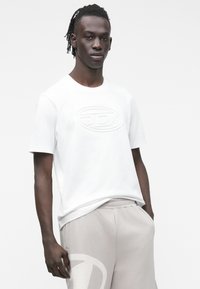 Vyras su trumpais dreadais dėvi baltą marškinėlį su reljefiniu logotipu ir šviesiai pilkas kelnes su dideliu išblukusiu logotipu, stovintis prieš baltą foną.