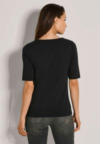 Vrouw met lang bruin haar in een eenvoudige zwarte T-shirt met korte mouwen en donkergrijze jeans, met haar rug naar de camera tegen een lichte muur.