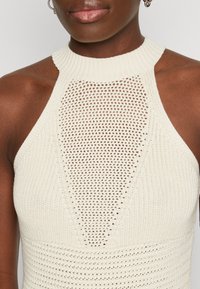 Top halter en tricot crème, présentant un motif pointelle au centre et des détails en côtes ; sans manches avec un col haut.