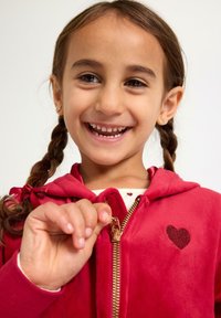 Rød hættetrøje med lynlås og hjertepatch; blød tekstur, foret hætte og guld hardware; båret over en hvid skjorte med røde hjerter.