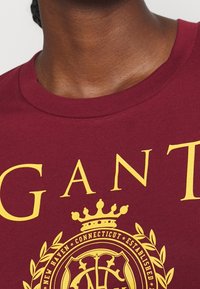 Bordeauxfarbiges Baumwoll-T-Shirt mit großem gelbem Text und einem runden Logo mit einer Krone und Lorbeerdesign, das sich auf der Vorderseite befindet.