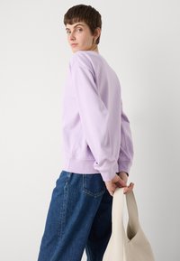 Lyse lilla genser med rund hals og lange ermer, ribbestrikkede mansjetter og nederst, kombinert med løstsittende mørkeblå jeans og en beige håndveske.