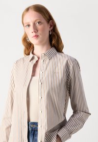 Camicia a righe beige e bianca, con chiusura a bottoni, colletto e maniche lunghe. Tessuto leggero, vestibilità casual, dettaglio del logo sul petto.