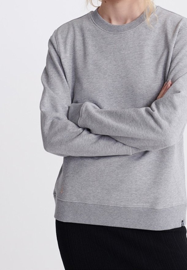 Personne portant un sweat-shirt gris uni à col rond avec manches longues, bras croisés, et une jupe noire sur un fond clair.