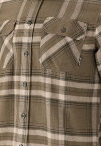 Bruin geruit flanellen shirt met donkergroene en roze strepen, knoopsluiting en twee borstzakken met kleppen. Getextureerde stof met een zachte afwerking.
