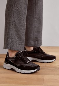 Black suede and mesh sneakers with a chunky white sole, featuring lace closure and metallic accents. Worn with gray herringbone trousers.