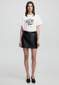 Camiseta blanca con gráfico en forma de corazón negro, combinada con una mini falda de cuero negra con costura frontal y trabillas, y zapatos negros de tacón.