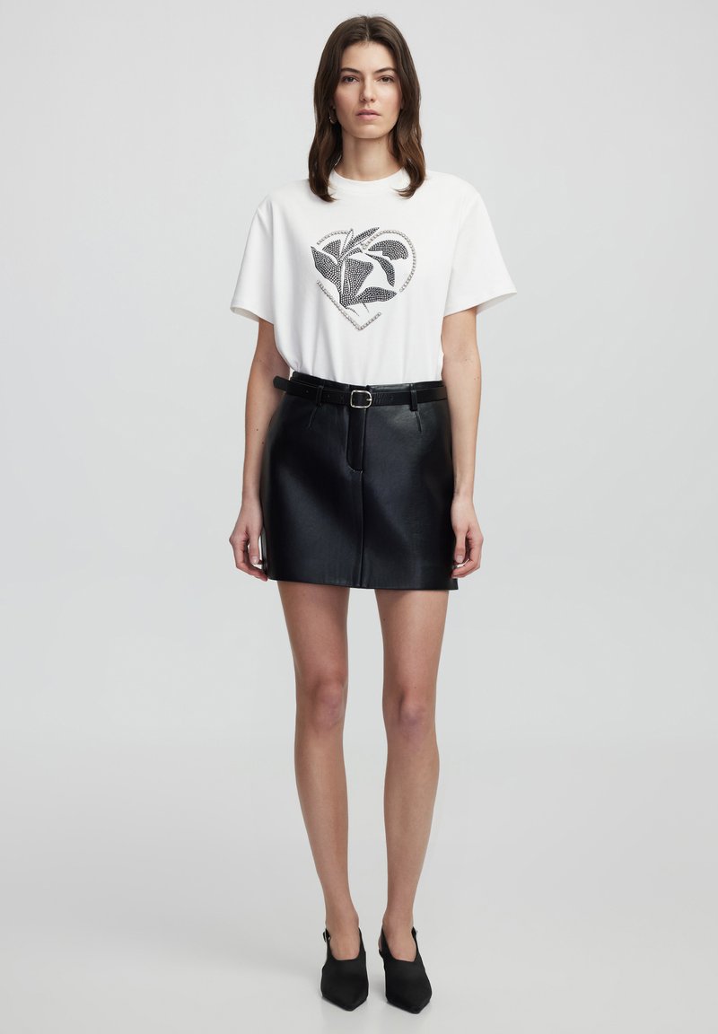 Witte t-shirt met een zwart hartvormig grafisch ontwerp, in combinatie met een zwarte leren mini-jurk met een voorkantnaad en riemlussen, en zwarte hoge hakken.