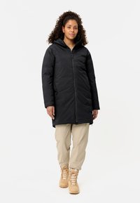 Parka nera isolata con zip frontale e cappuccio, caratterizzata da una texture liscia, maniche lunghe e lunghezza fino a metà coscia. Indossata con pantaloni beige e stivali marroni.